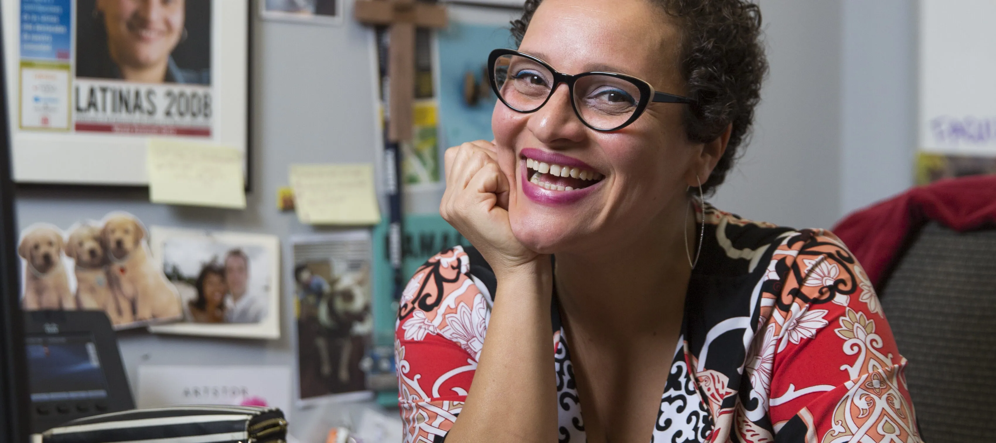 A olive-skinned woman smiles brightly, seemingly mid-laugh, as she sits at an office desk, her right hand placed beneath her chin. Her brown curly hair is worn in a cropped pixie cut and she wears cat-eye glasses. In front of her is a closed Apple laptop; the wall behind her holds two framed certificates or degrees, a framed newspaper, a wooden cross, and other pasted photos.