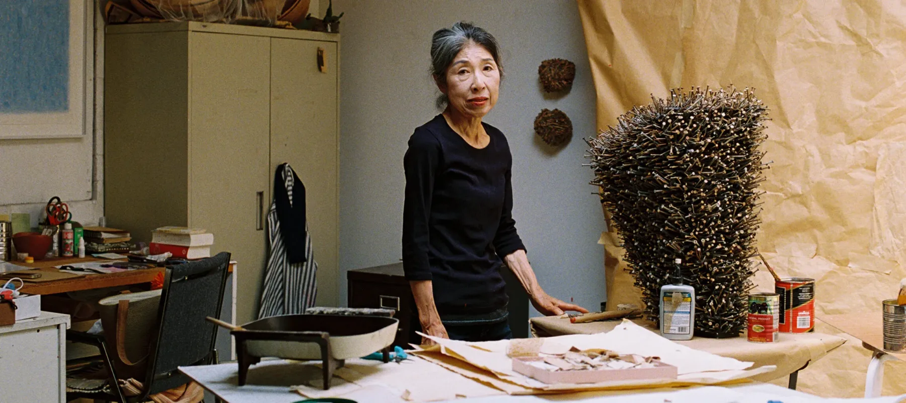 In a white brick artist's studio, a small light-skinned Japanese woman stands amongst various supplies, including work paper, paper sculptures, scissors, glue sticks, and paint cans. She wears a black shirt and stares directly at the camera, unsmiling. Her black hair is pulled back and she has streaks of white hair at her hairline's part.