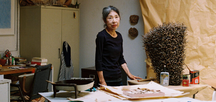 In a white brick artist's studio, a small light-skinned Japanese woman stands amongst various supplies, including work paper, paper sculptures, scissors, glue sticks, and paint cans. She wears a black shirt and stares directly at the camera, unsmiling. Her black hair is pulled back and she has streaks of white hair at her hairline's part.