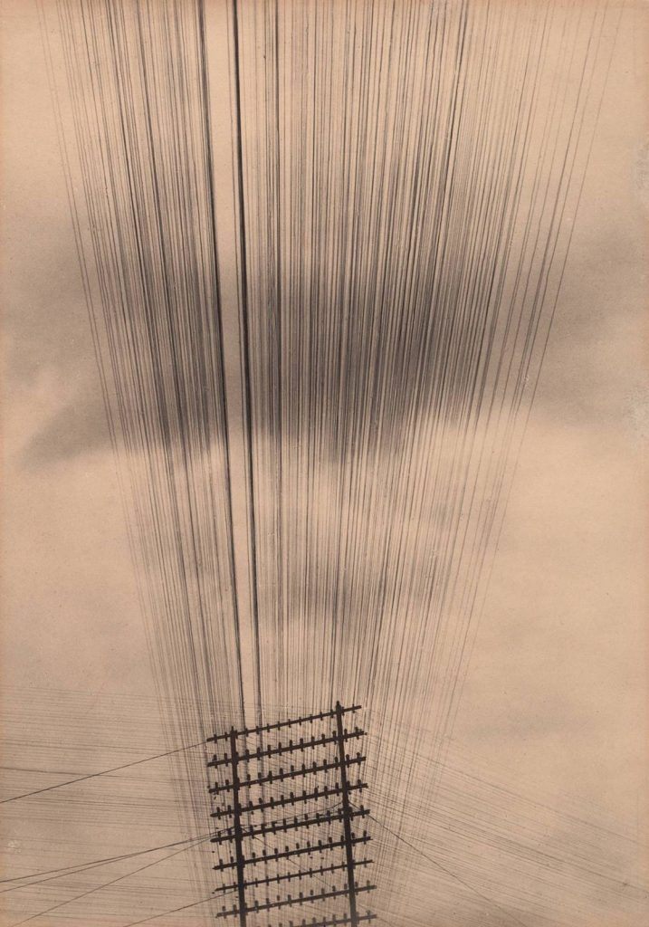 A sepia-toned photo shows a power line tower at the photo's bottom with many thin, straight power lines emanating from it and crossing a cloudy sky, all taking up the upper two-thirds of the photo.