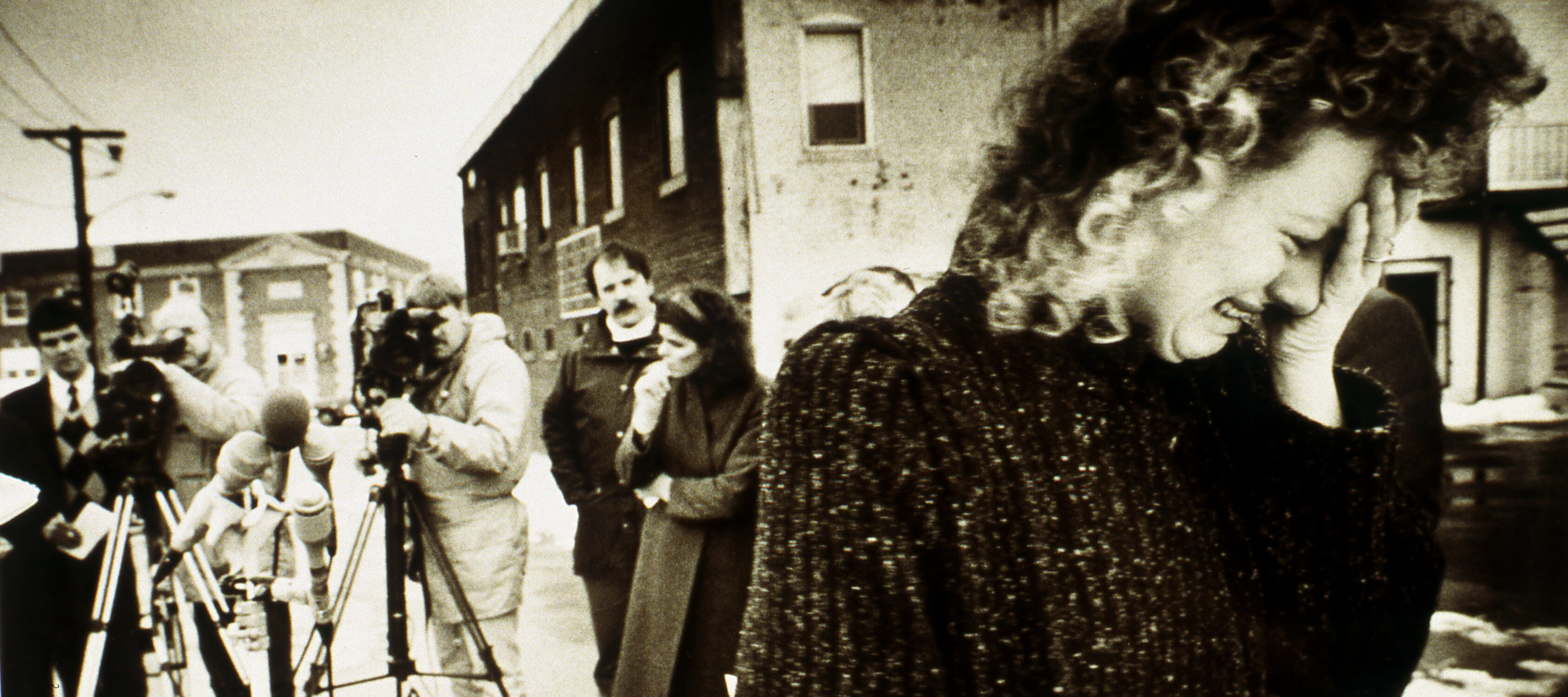 A black-and-white photograph of a light-skinned adult with curly hair crying into their hand. They wear a sweater and stand in the foreground, seen waist up, while several photographers swarm in the background behind a microphone stand set up in the middle of the street.
