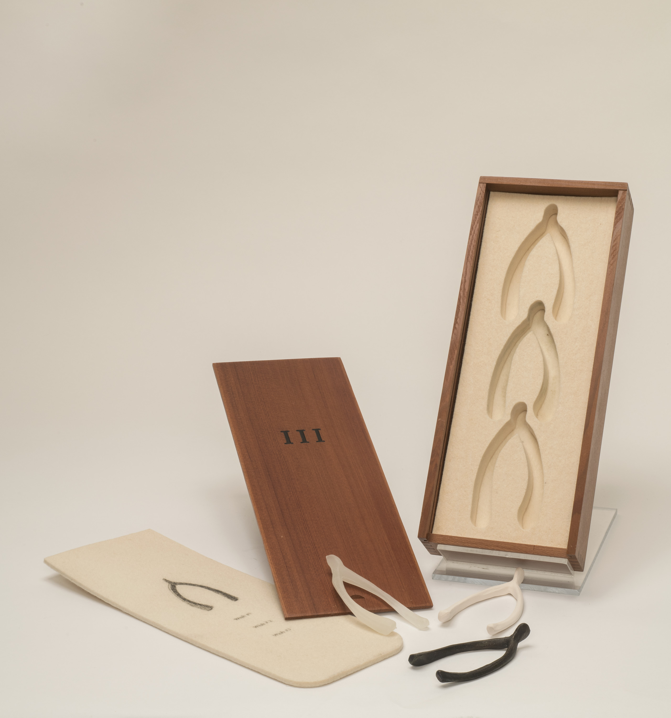 A box with indentations for three wishbones is propped up with it's wooden cover next to it with the symbol "III" printed in black ink. Next to it is a paper with a wishbone printed on it, and three physical wishbones, seemingly from the box. Two are white, one is black.