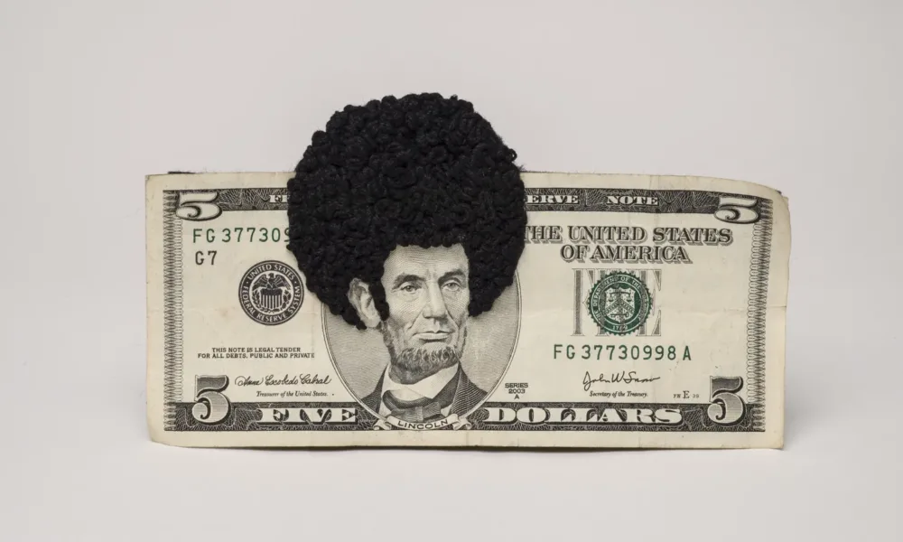 U.S. five-dollar bill has an embroidered afro and sideburns stitched onto the portrait of Lincoln’s head. One-third of the afro protrudes beyond the top of the bill.