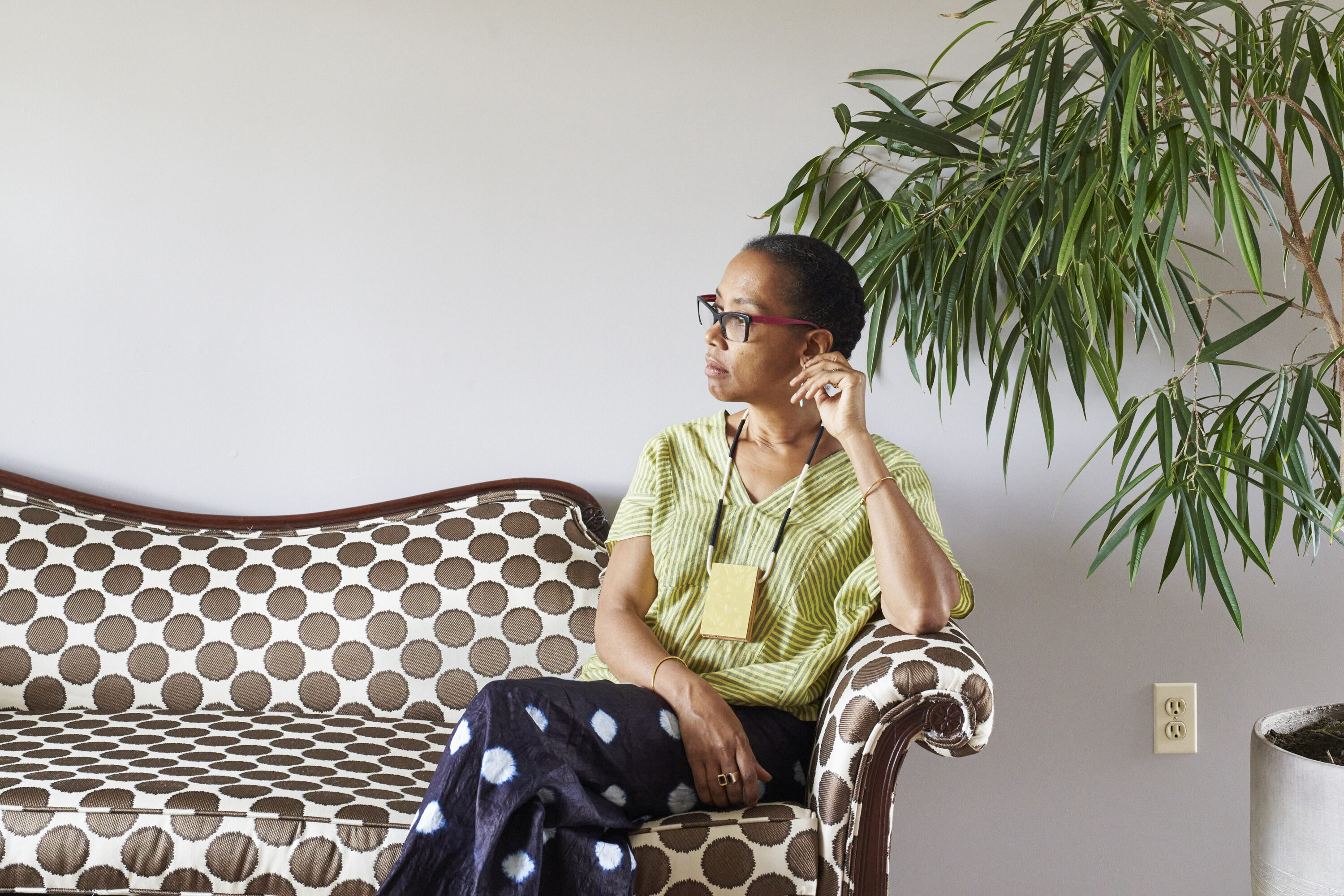 Sonya Clark calmly sits on a polka-dot couch against a white wall, next to a large plant. She is a medium-skinned adult woman with dark hair pulled back, looking to her right with her legs crossed. She wears a striped top, black pants with large white dots, glasses, and a necklace.