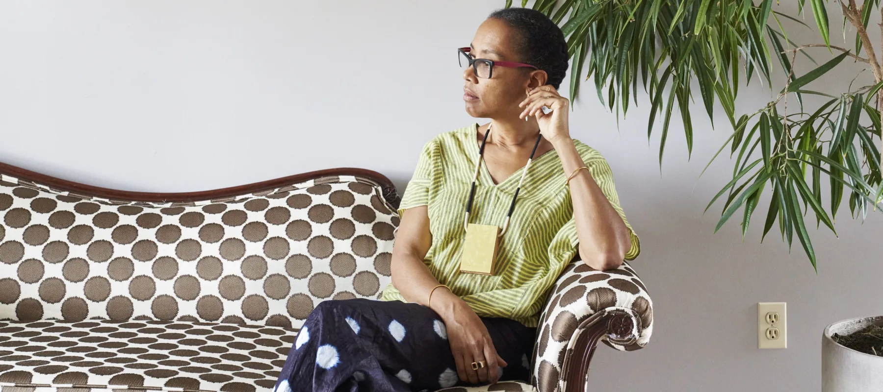 Sonya Clark calmly sits on a polka-dot couch against a white wall, next to a large plant. She is a medium-skinned adult woman with dark hair pulled back, looking to her right with her legs crossed. She wears a striped top, black pants with large white dots, glasses, and a necklace.