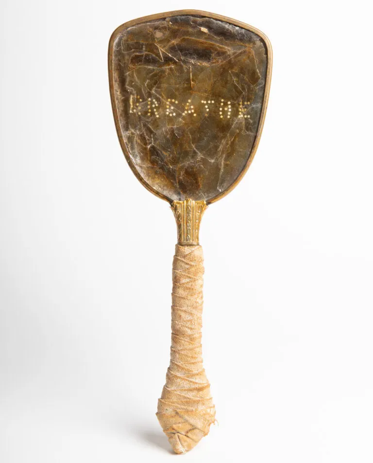 An antique hand mirror against a white background. The handle of the mirror is wrapped in light colored cloth. The head of the mirror is a brown, non-reflective material. The word “breathe” has been perforated onto the surface.