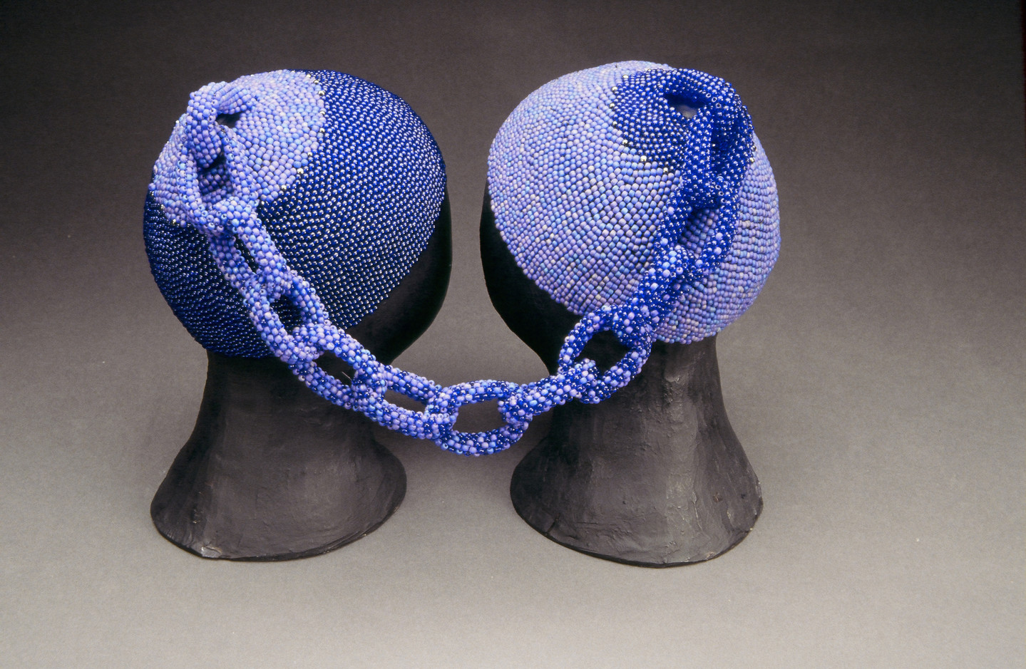 Two head caps made of small, blue glass beads rest on two black mannequin heads. The two caps are connected at the tops by a beaded chain. The left cap is made of darker blue beads and the right cap is made of lighter blue beads. The chain combines both shades.