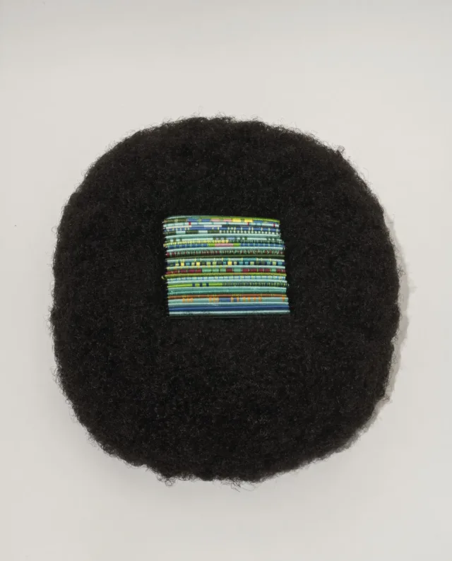 A square of bright, multicolored threads of blue and green tones rest in the center of a circular afro wig made of black hair.