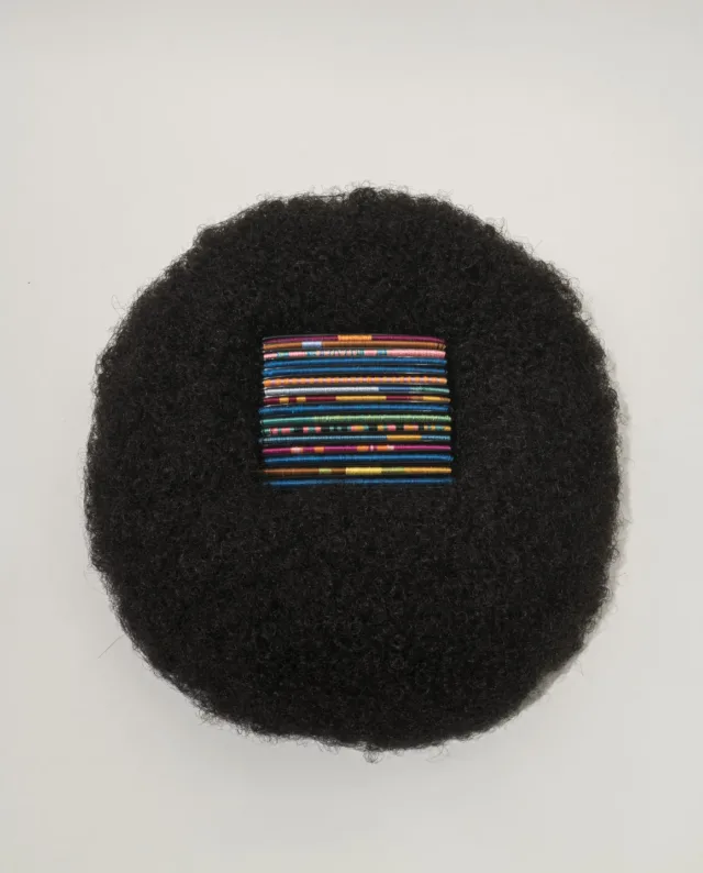 A square of bright, multicolored threads rest in the center of a circular afro wig made of black hair.