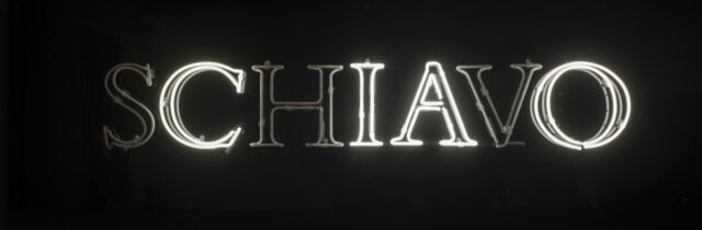 White, capital, neon letters spell the Italian word 'schiavo' in serif text against a black background. The 's,' 'h,' and 'v' are dark, leaving the letters to spell 'ciao.'