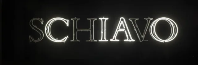 White, capital, neon letters spell the Italian word 'schiavo' in serif text against a black background. The 's,' 'h,' and 'v' are dark, leaving the letters to spell 'ciao.'