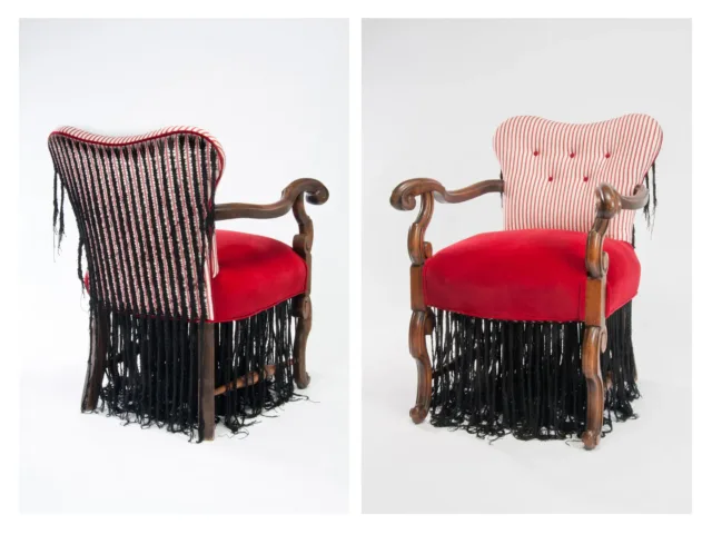 A sculpture of an arm chair with wooden legs and arms and a red fabric seat. Black cotton thread has been stitched onto the back and underside of the chair in the form of braided cornrows. The braids hand down beneath the chair, dangling to the ground.