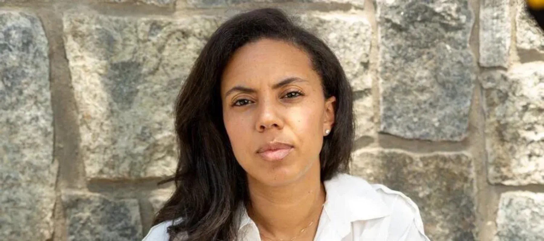 A medium-skinned woman with straight black hair sits against a stone wall wearing a flowy white dress with a collar and belt. Her hands are on her knees and she looks at the camera with a slightly furrowed brow.