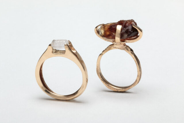 Two rings with gold bands and small gem-like forms on top. The gems are crafted from raw sugar. The left ring is perfectly circular with a small, smooth, white sugar crystal on top. The right ring is a slightly rougher circle, with a larger, lumpy, brown sugar crystal on top.