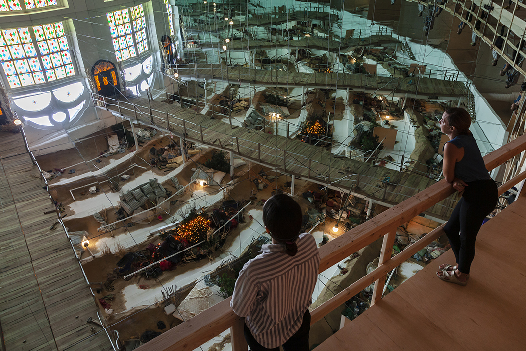 A fish-eye view of a large art gallery space that features sand dunes riddled with random household objects; a wall of floor-to-ceiling mirrors, and imitation church stained glass hung or projected onto white fabric. Two women stand on a wooden balcony in the lower right of the frame looking down on the scene.