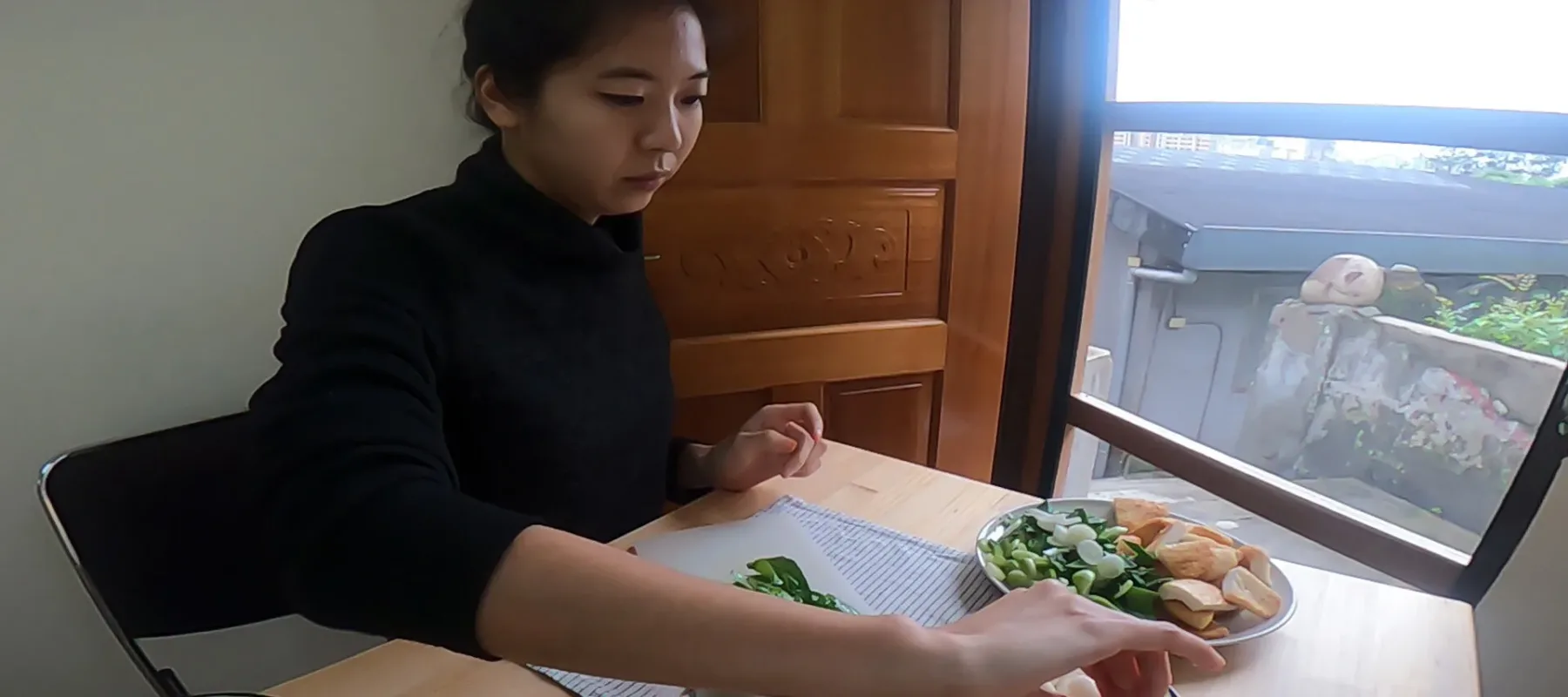 A light-skinned adult woman wearing a black turtleneck with sleeves rolled up to her elbows and her black hair pulled back sits at a wooden table by a window. She is making ddukbokki by slicing greens and preparing white rice paddies shaped like small sausages.