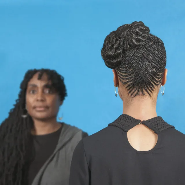 A color photograph featuring two dark skinned women in front of a blue background. The woman on the left has long brown hair pulled over her right shoulder. She is facing the camera and wears a black shirt with a grey cardigan. The woman on the right stands with her back to the camera wearing a black shirt with a single button at the top. Her brown hair is braided in an elaborate pattern and pulled up on the left side of her head.