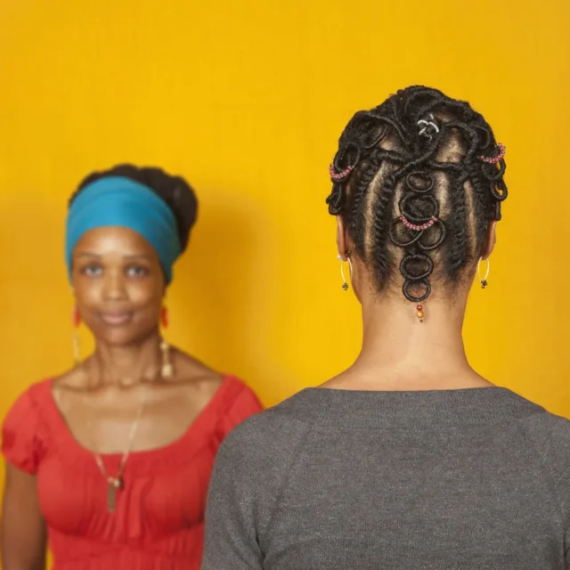 Two medium-dark skinned adult women stand in front of a goldenrod background. The woman on the left faces the viewer and wears a teal head wrap and a red top. The woman on the right faces away from the viewer, showcasing her dark hair braided into an elaborate pattern with colorful beads.