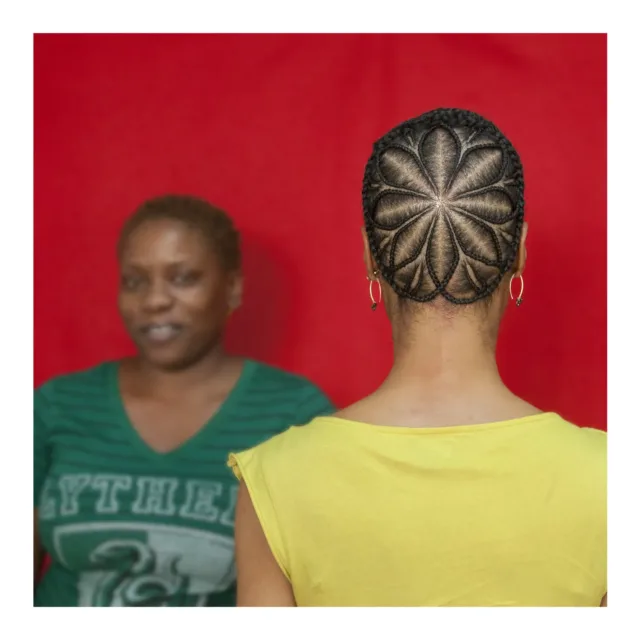 A color photograph featuring two dark skinned women in front of a red background. The woman on the left has very short brown hair and faces the camera, she is wearing a green v-neck t-shirt. The woman on the right stands with her back to the camera wearing a yellow shirt. Her brown hair is braided in an elaborate star pattern.