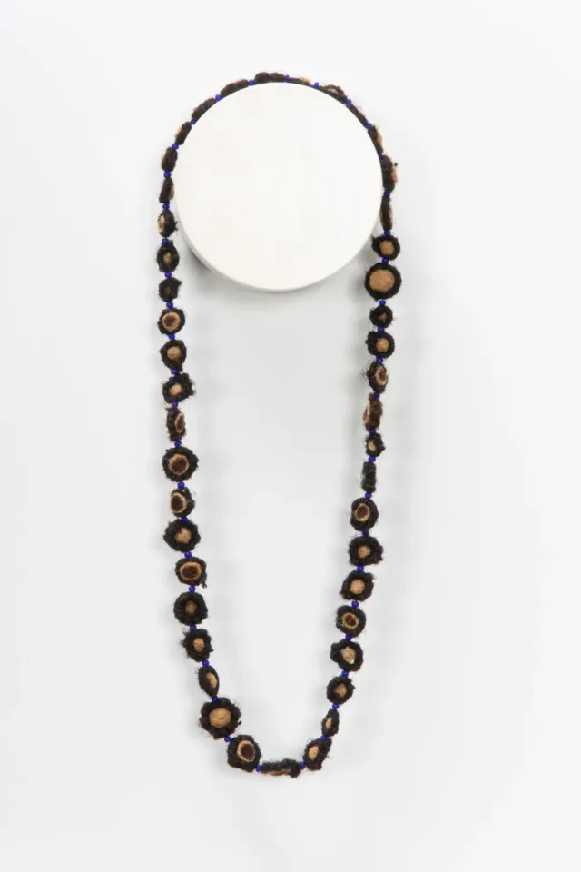 A necklace made from coiled, balled, and twisted dark human hair hangs on the wall. The hair is wrapped around small beads.