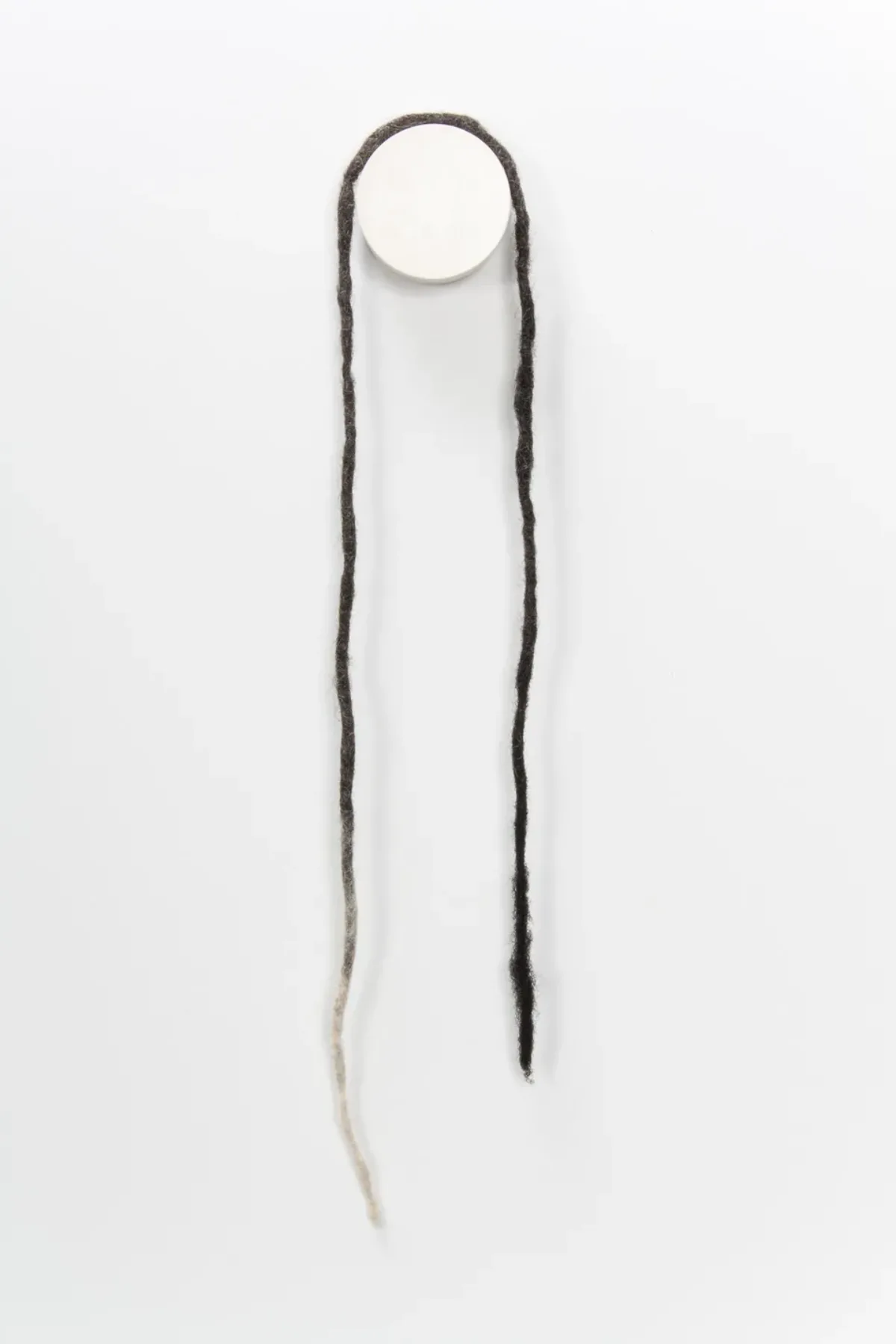A necklace made from coiled, balled, and twisted dark human hair hangs on the wall. The necklace is formed from a long, open-ended dreadlock that becomes white at its end.