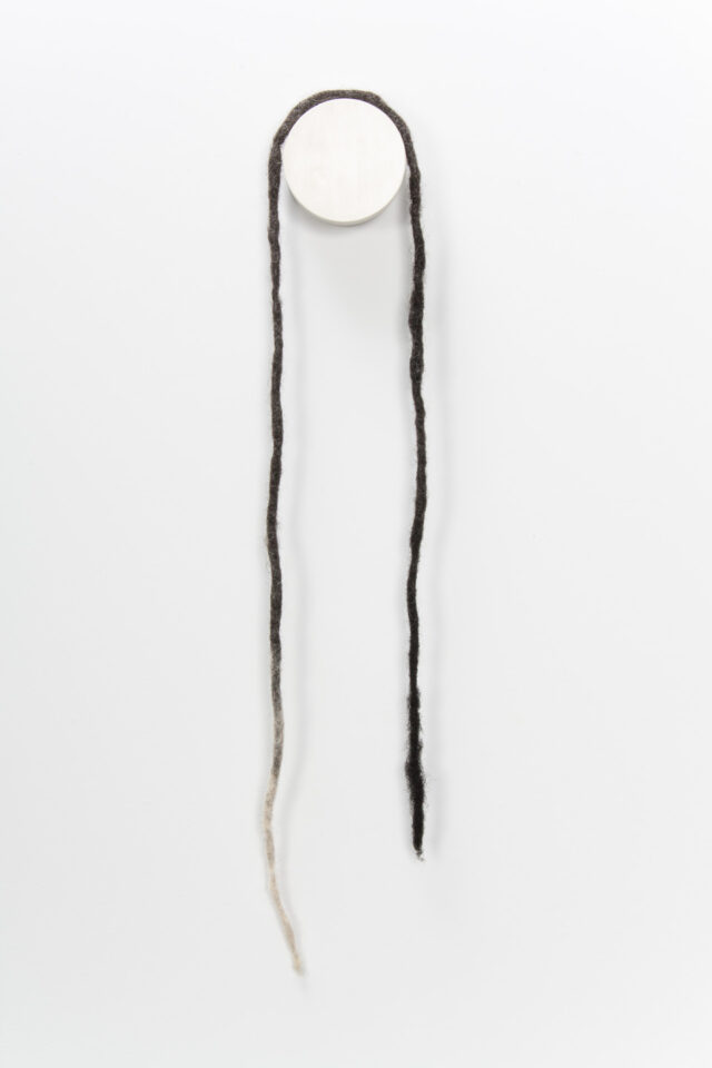 A necklace made from coiled, balled, and twisted dark human hair hangs on the wall. The necklace is formed from a long, open-ended dreadlock that becomes white at its end.