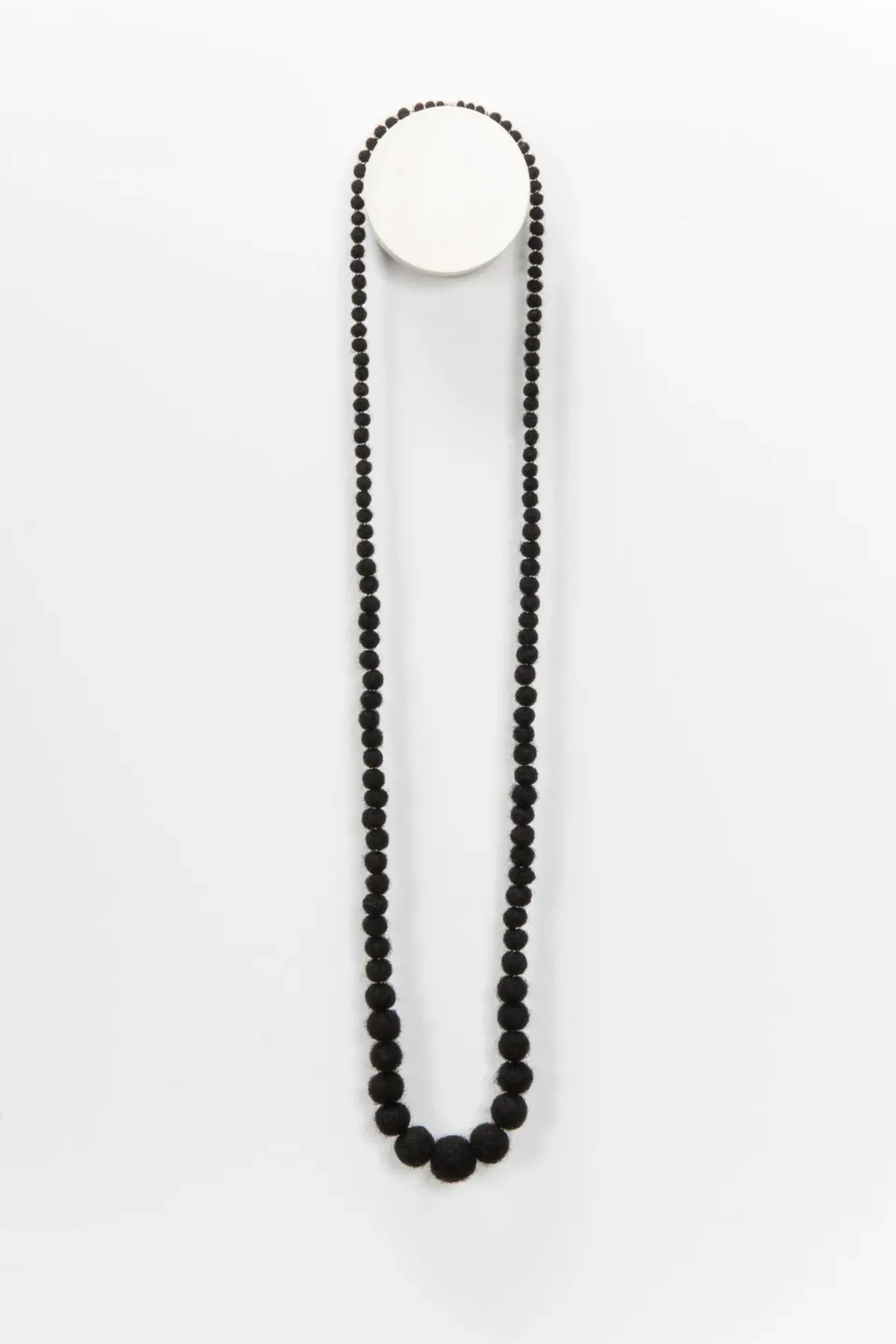 A necklace made from coiled, balled, and twisted dark human hair hangs on the wall. The hair is rolled into balls that grow in size toward the middle.