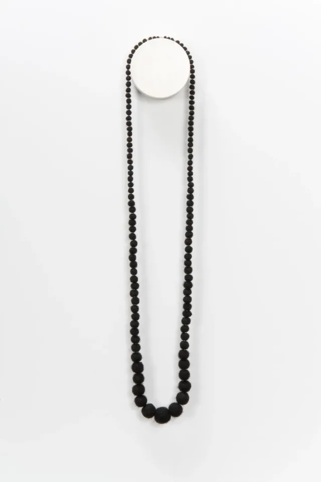 A necklace made from coiled, balled, and twisted dark human hair hangs on the wall. The hair is rolled into balls that grow in size toward the middle.