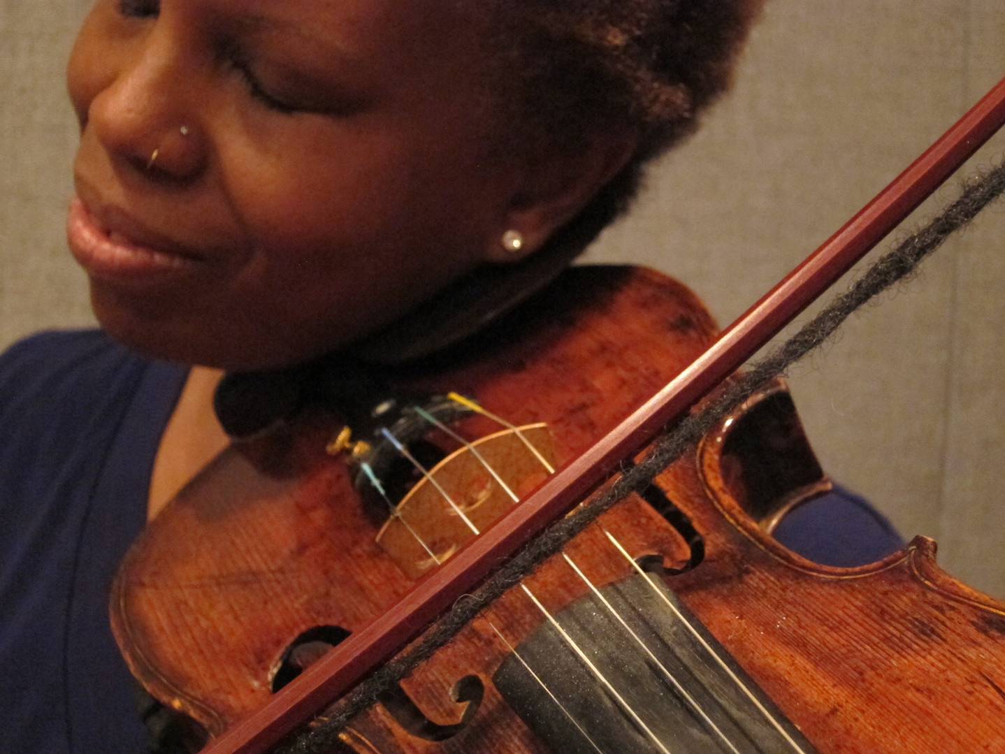 A color photograph of a dark-skinned woman holding a violin and playing its strings with the bow strung from a dreadlock.