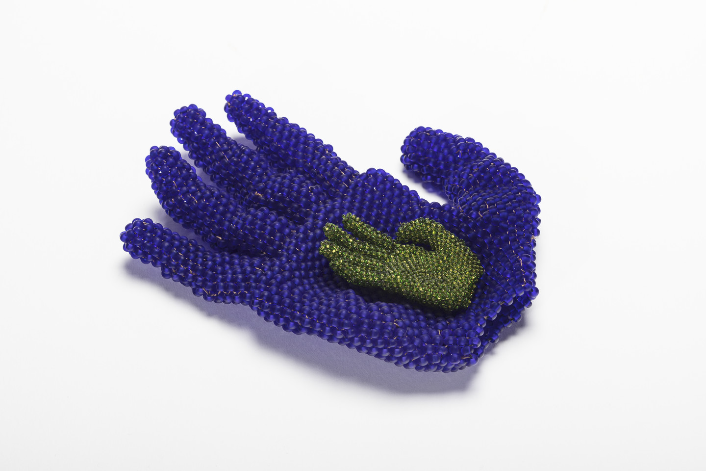 A beaded sculpture of two hands. A larger hand, made of blue glass beads, rests palm up. It cradles a smaller hand made of green glass beads, which is centered in the same position in the larger hand’s palm.