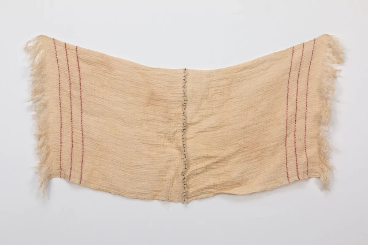 A rectangular, light colored sheet of fabric with three vertical red stripes on either end. The cloth drapes in the center, where two halves have been sewn together using black thread.