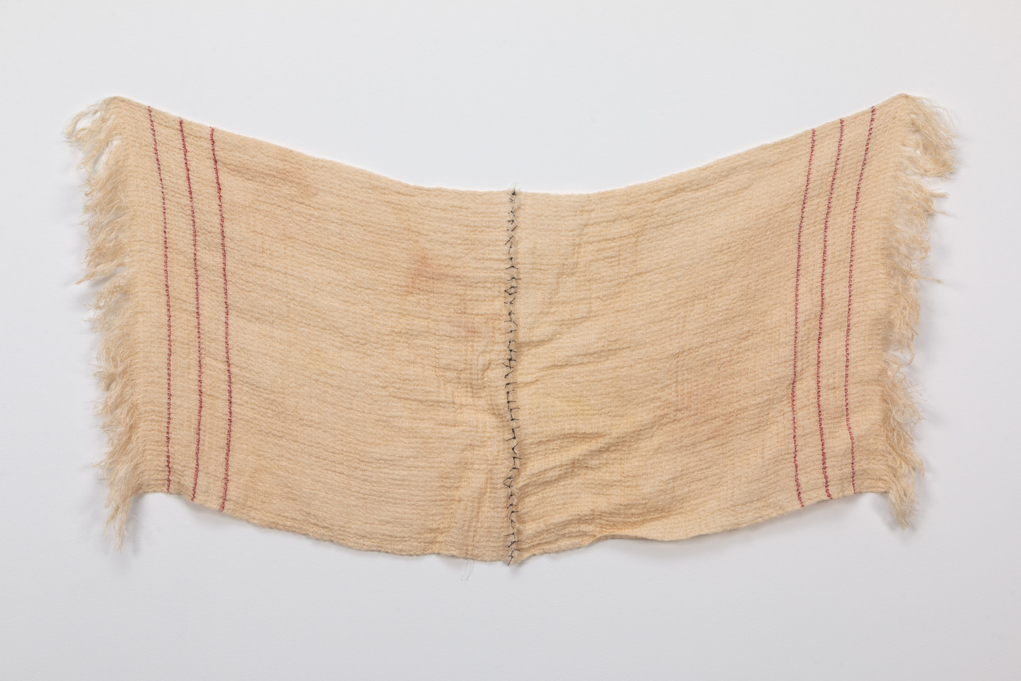 A rectangular, light colored sheet of fabric with three vertical red stripes on either end. The cloth drapes in the center, where two halves have been sewn together using black thread.