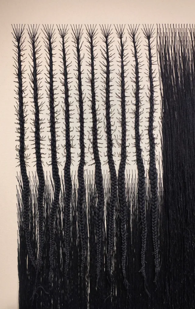 Detail view of a vertical rectangular canvas woven with black cotton thread. The thread has been braided into the approximate form of an American flag. Where the stars would be, the thread forms branching cornrows, and where the stripes would be, the thread hangs densely, like hair.