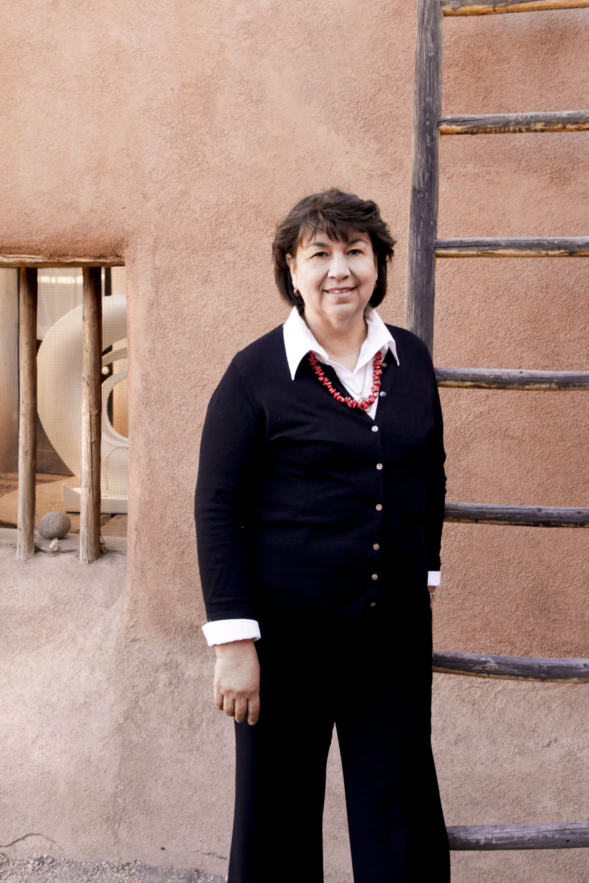 A light skinned woman with short brown hair wearing a black sweater, black pants, a white collared shirt, and a coral necklace, stands smiling in front of a ladder resting against a light pink stucco building. You can see a round white sculpture sitting on a table through the window of the building.