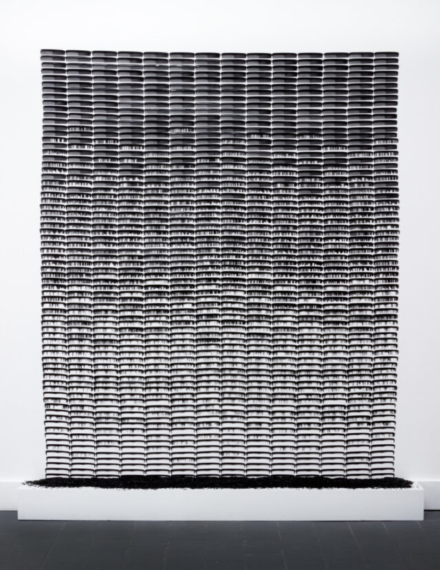 A large rectangular sculpture made of black, plastic pocket comes attached side by side in rows against a white background. As the combs descend, more and more of their plastic teeth are removed, leaving gaps and open spaces. The removed pieces of comb teeth are pooled at the base of the sculpture.
