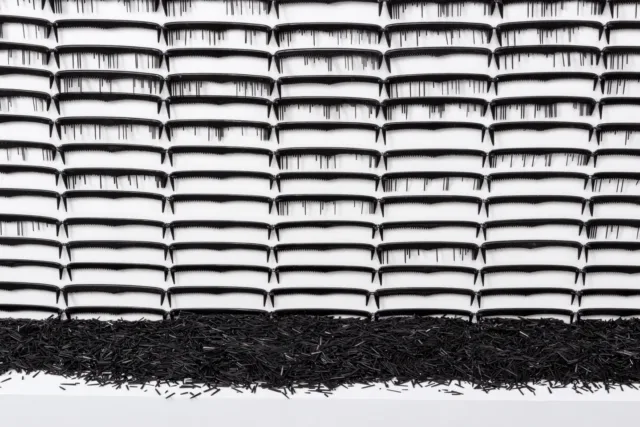 Detail image of a large rectangular sculpture made of black, plastic pocket comes attached side by side in rows against a white background. As the combs descend, more and more of their plastic teeth are removed, leaving gaps and open spaces. The removed pieces of comb teeth are pooled at the base of the sculpture.