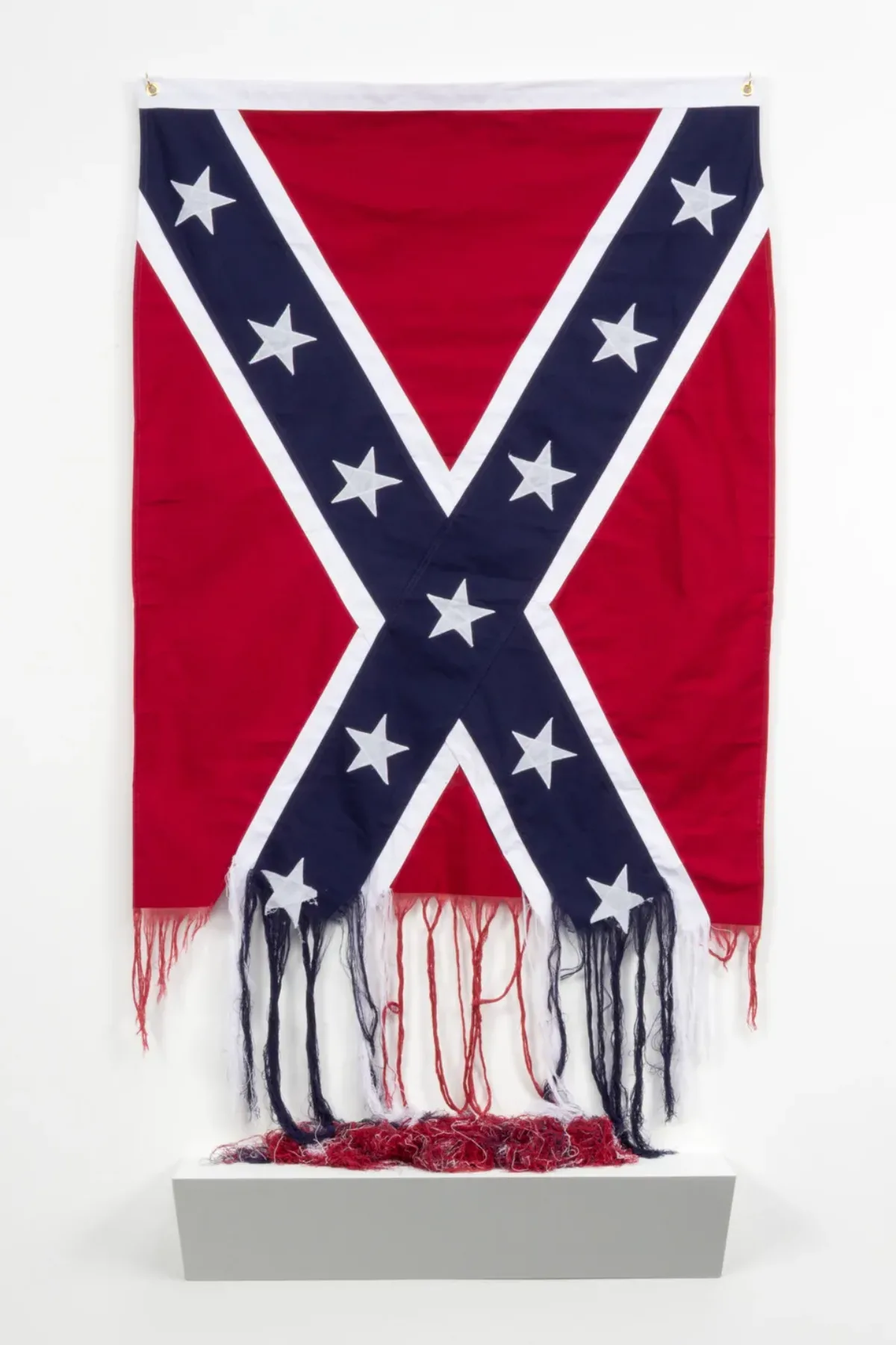 An image of an American Confederate Battle Flag, which is partially unraveled at the bottom. The unraveled threads hang down loosely and pool into a pile on a white platform beneath the flag.