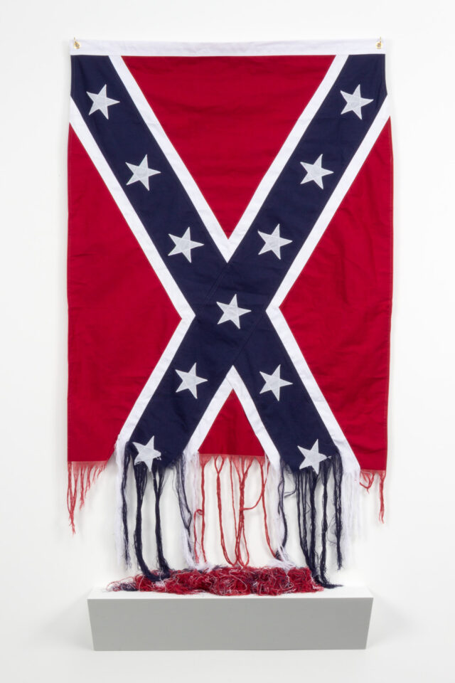 An image of an American Confederate Battle Flag, which is partially unraveled at the bottom. The unraveled threads hang down loosely and pool into a pile on a white platform beneath the flag.
