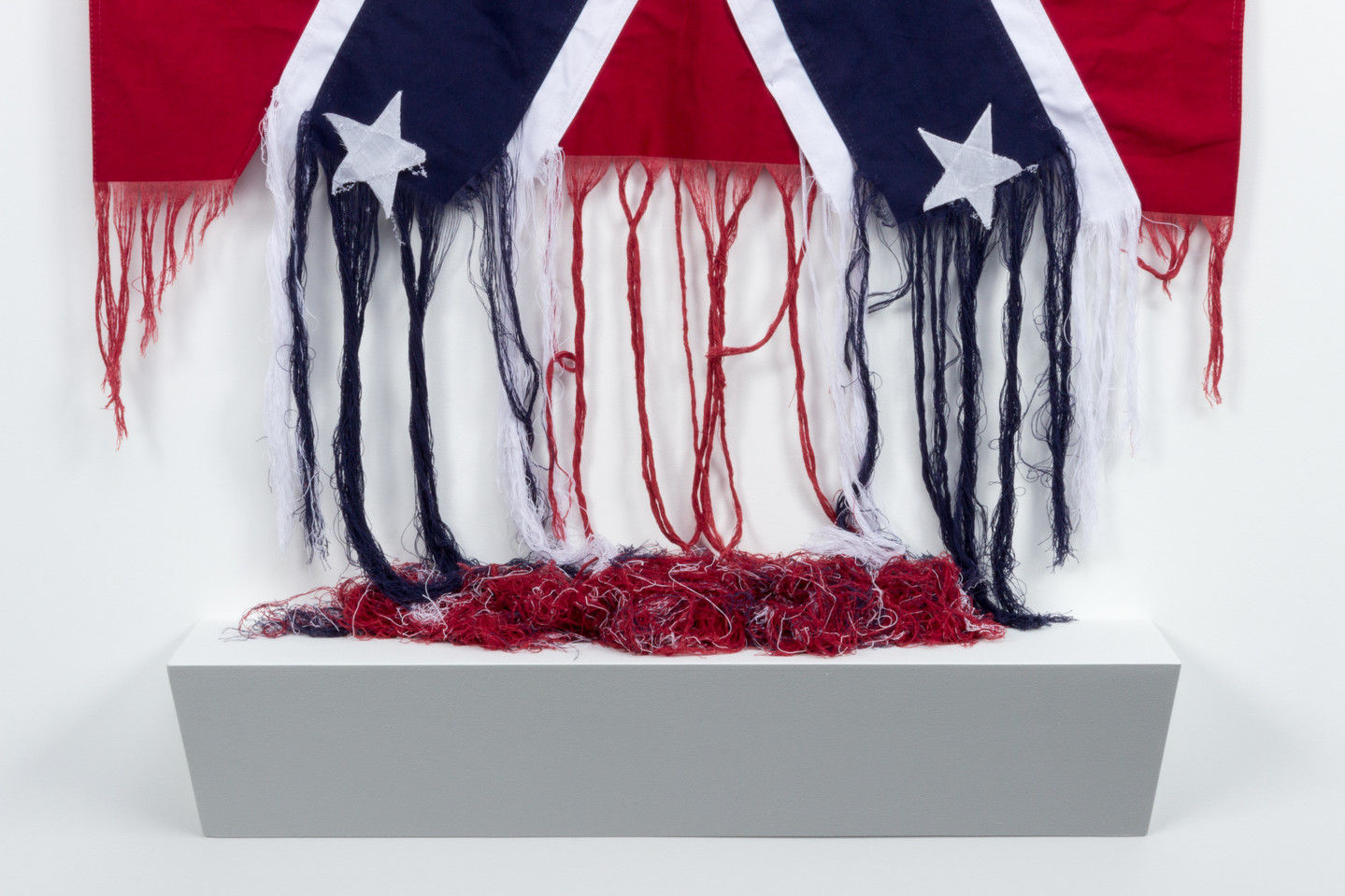 The bottom of an American Confederate battle flag, which has been unraveled. The red, white, and blue threads hang loose, and pool onto a shelf below.