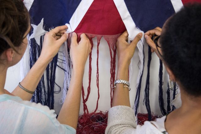 A photograph of two figures standing side by side, unraveling the threads of an American Confederate battle flag. The figure on the left has light skin, and the figure on the right has darker skin. They face away from the camera, with their hands in the center of the image pulling loose the threads.
