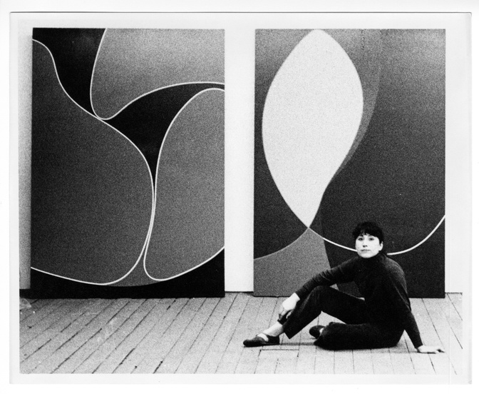 A black-and-white photo of a light-skinned woman sitting on the wooden floor in front of two large abstract works featuring curved lines and soft shapes. She has bangs and wears her hair pulled back and wears a dark turtle neck and dark pants.