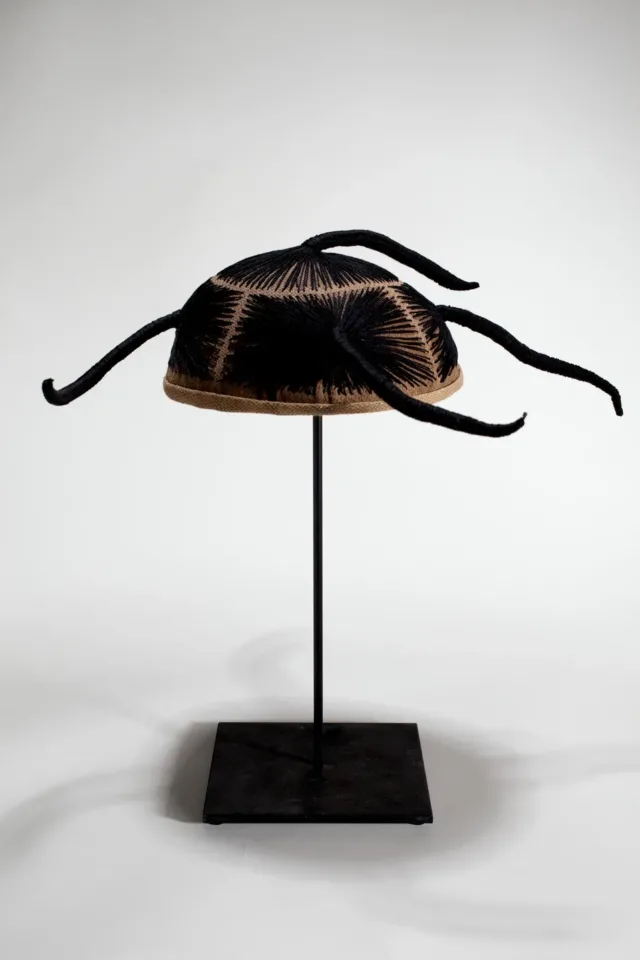 A wig cap sits mounted on a black stand. Black thread is stitched into the cap, mimicking hair. The thread forms five finger-like forms that protrude from all sides.