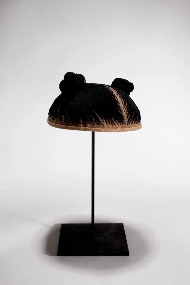 A wig cap sits mounted on a black stand. Black thread is stitched into the cap, mimicking hair. The thread forms two small buns.