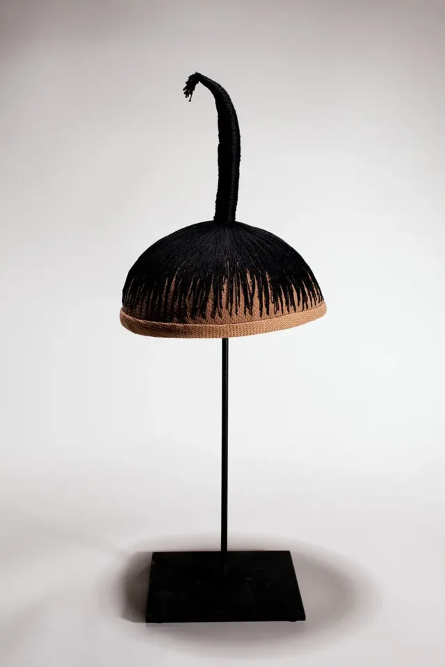 A wig cap sits mounted on a black stand. Black thread is stitched into the cap, mimicking hair. The thread is stitched into a single column, sticking up vertically from the cap.