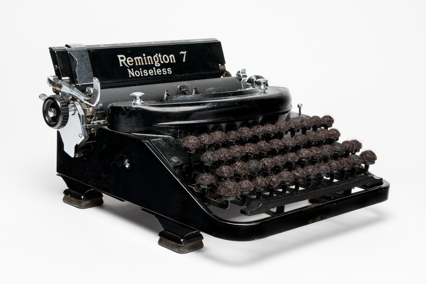 An antique, black and silver typewriter with the words “Remington 7 Noiseless” emblazoned on the top. The lettered keys of the typewriter have been replaced with balls of dark brown hair.