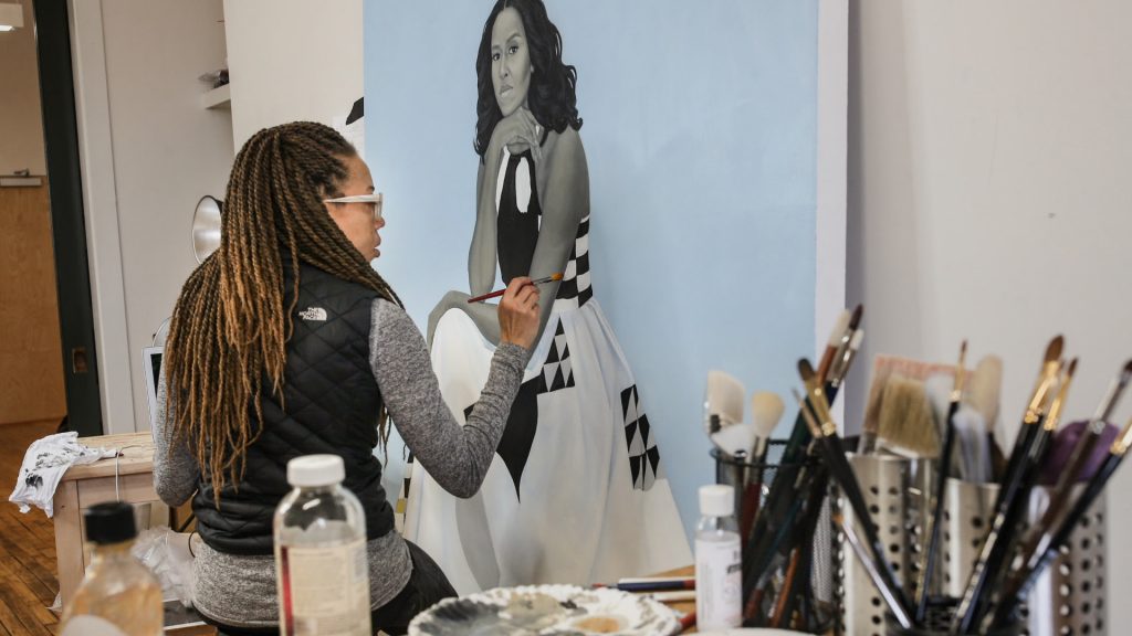 An African American woman with long braids is at work on a painting of Michelle Obama, presented in grey-scale with a powder blue background.