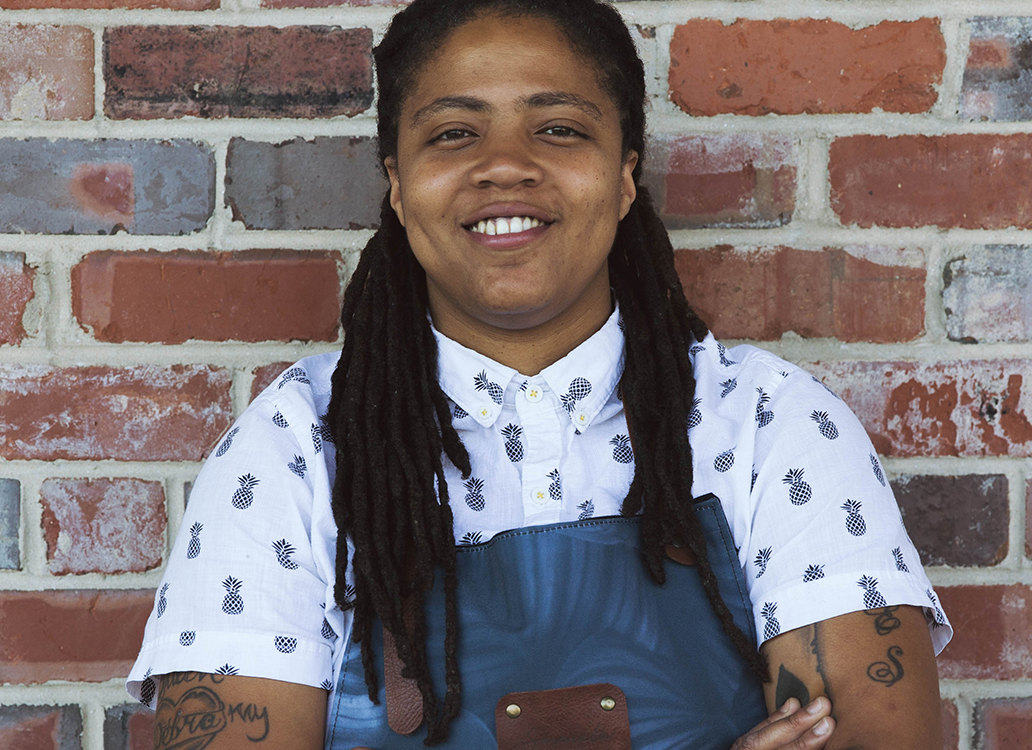 A dark skinned woman with long dreadlocks wearing a short-sleeved white collared shirt with a black pineapple pattern and a grey-blue apron stands in front of a brick wall. The woman is smiling and has tattoos on her arms just below the sleeves of her shirt