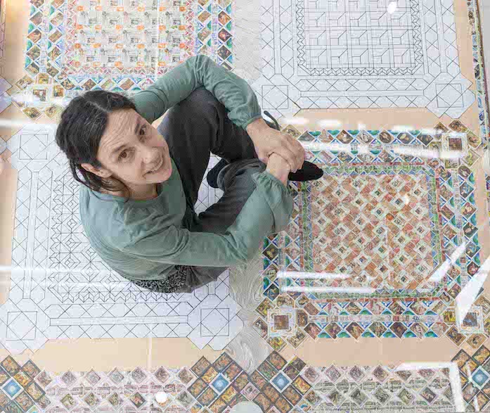 A light-skinned woman sits atop a colorful mosaic floor with her knees drawn up to her chest and her hands clasped on top of them. She looks up at the camera and smiles.