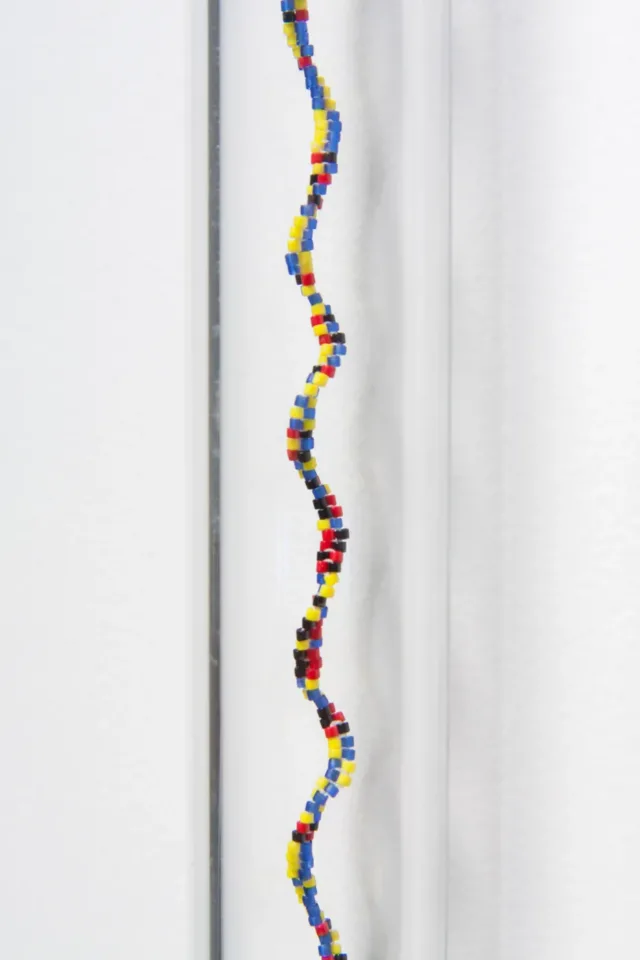 Detail image of a sculpture of a glass tube affixed to a white wall with a spiraling column of small, multicolored glass beads. The beads are blue, red, yellow, green and black.