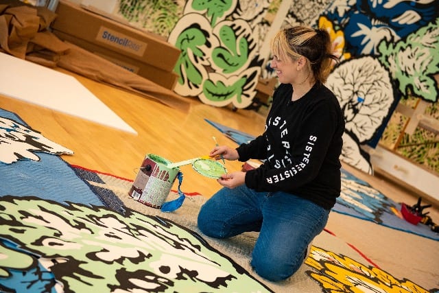 A light-skinned woman with black and blonde hair and bangs kneels on the floor atop large colorful designs. She smiles and her hands hold a small paintbrush and wooden paint stick over a can of mint green paint. There are multiple other colorful large-scale paintings of leaves and organic matter behind her.